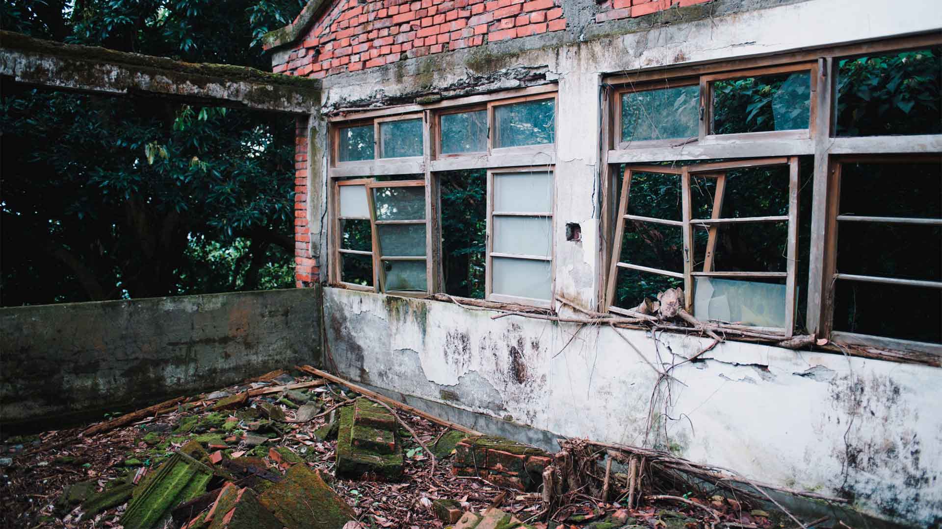 abandoned-old-building-with-destroyed-windows-forest Abandoned white building with red brick roof and concrete archway, broken windows and debris on the ground in a wooded area.
