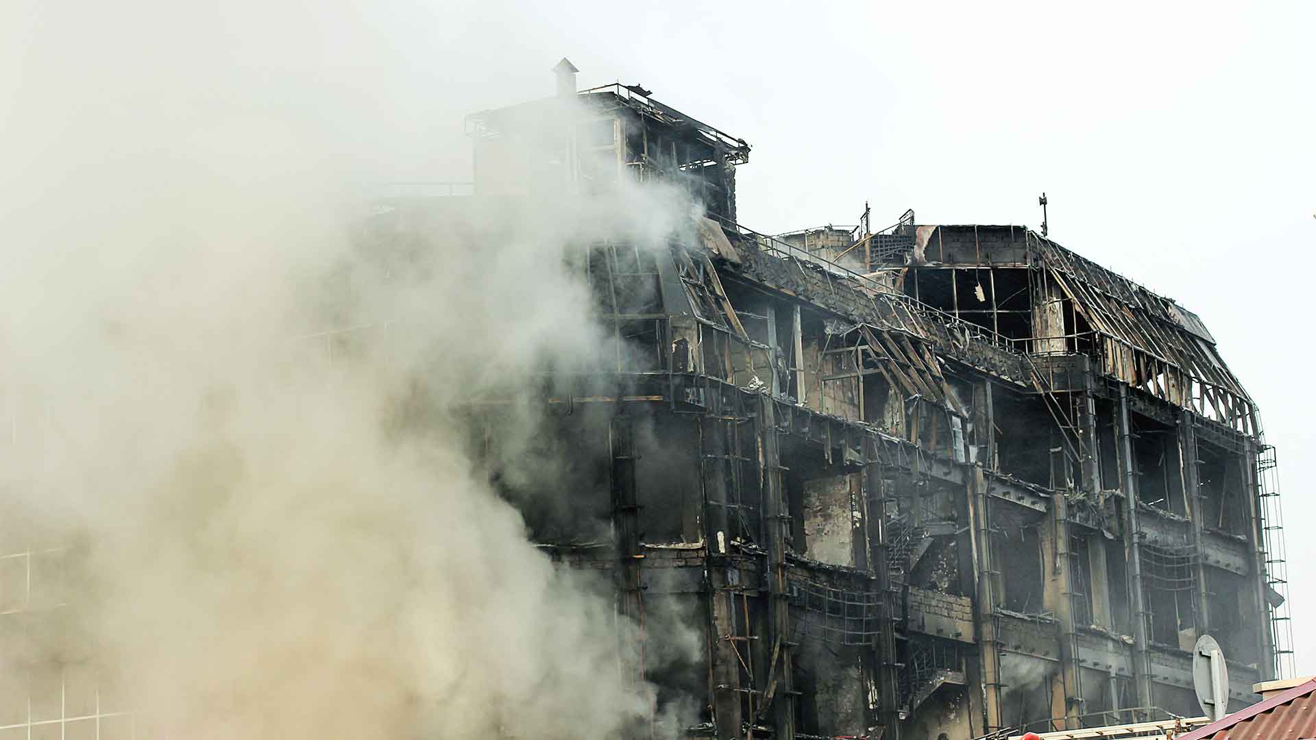 burning-shopping-center-mall-with-smoke a multi-story building on fire, covered in scaffolding, with a large plume of smoke rising from the blackened and charred structure against a gray and overcast sky.