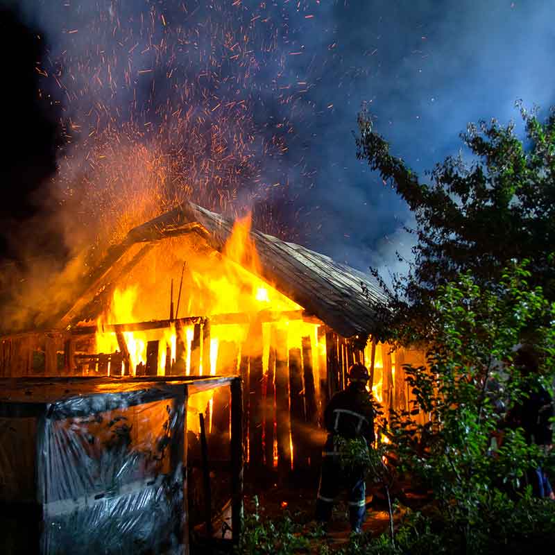 fire Firefighter extinguishing a house fire at night with water hose, wooden house with thatched roof ablaze with orange and red flames, dark sky filled with smoke.