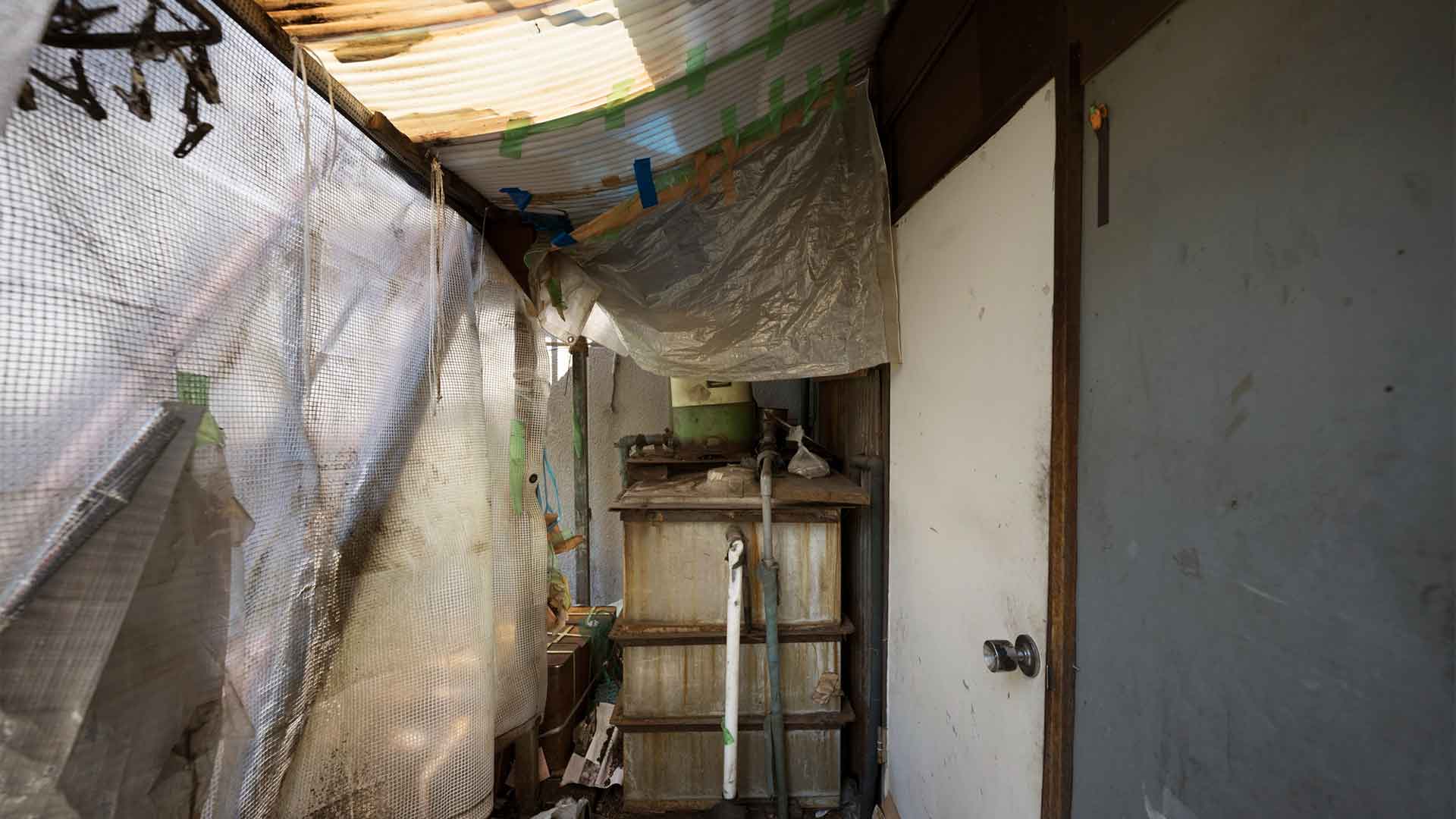 haunted-house-with-old-furniture-background Cluttered and dilapidated room with corrugated metal roof, concrete walls, wooden shelf, and white door.
