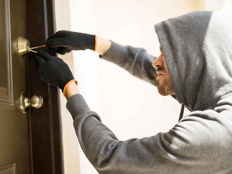 profile-view-man-with-hoodie-trying-pick-lock-house-forcing-his-entry person in gray hoodie and black gloves picking lock on wooden door with brass doorknob and deadbolt.